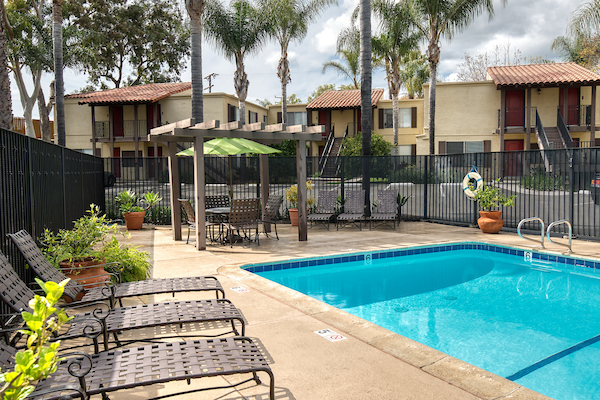 Outdoor swimming pool area with pergola, lounge seating and trees in background.